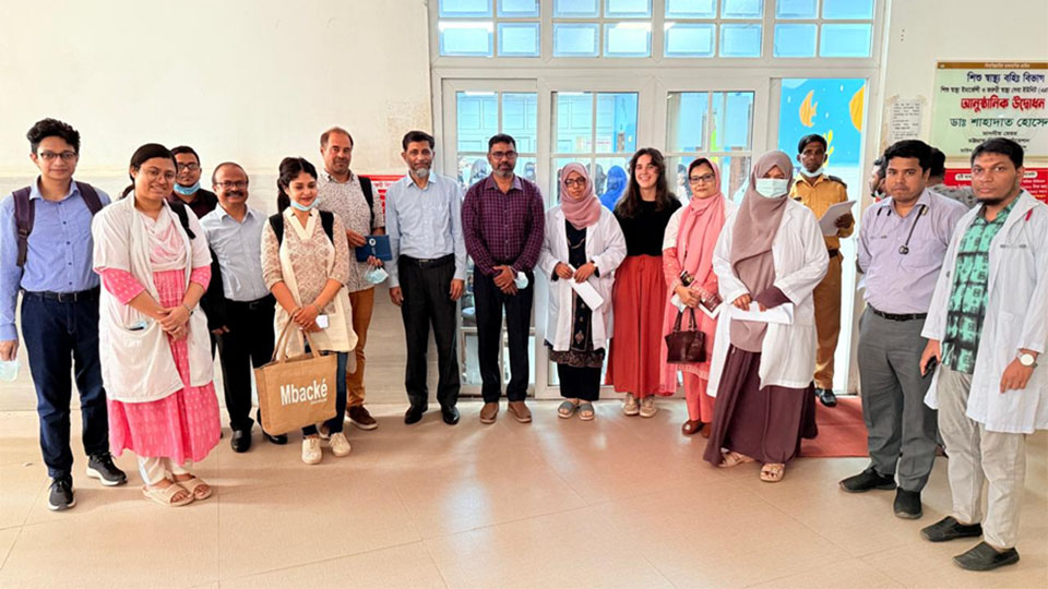 Dans un hall d'hopital, un groupe de personne pose, en arrière plan une entrée vitrifiée indique les Urgences pédiatriques en anglais et bangladais.